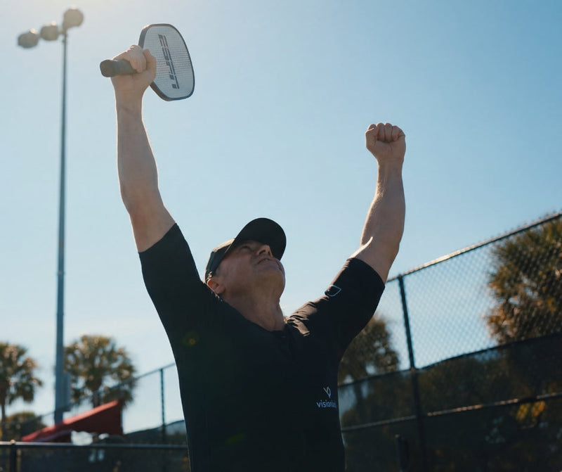 Man wearing a Visionbody EMS suit celebrating with a pickleball paddle on an outdoor court.